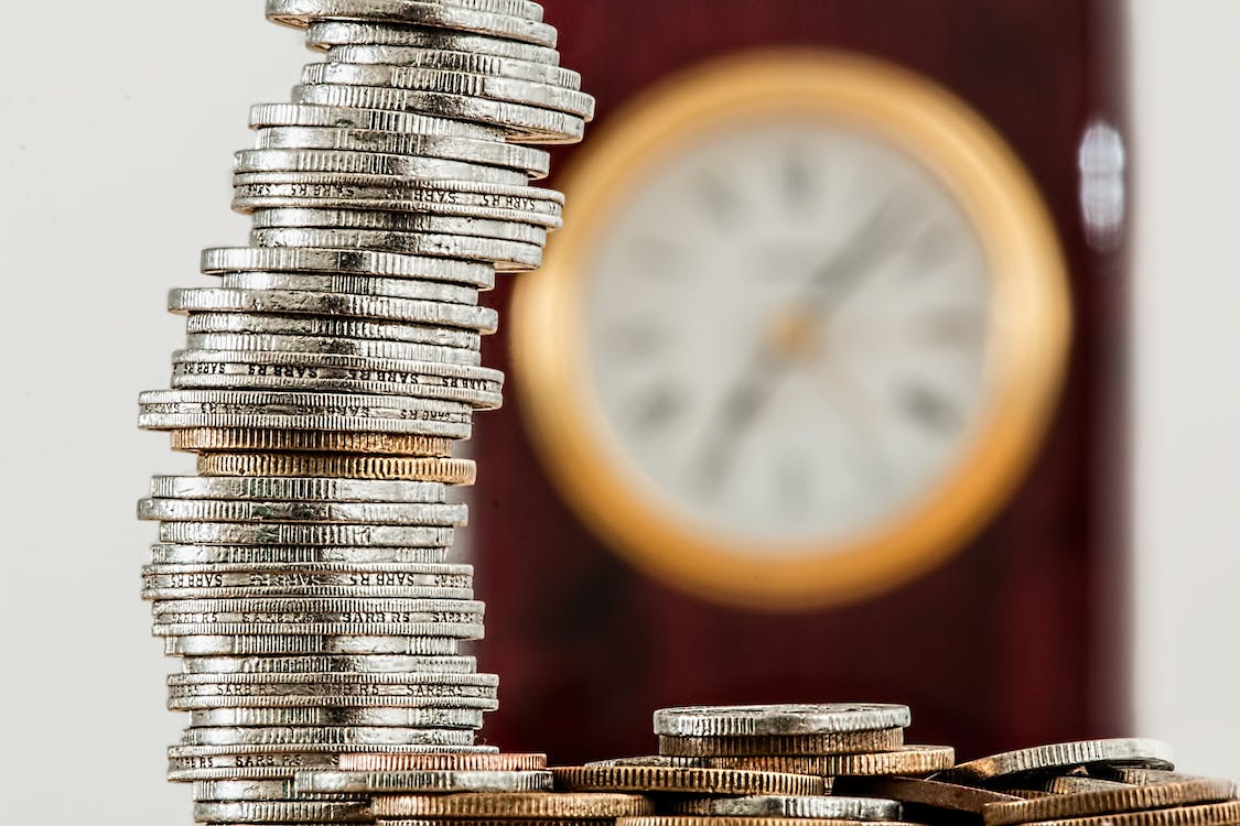 A selective focus photo of silver coins stacked in front of a clock.